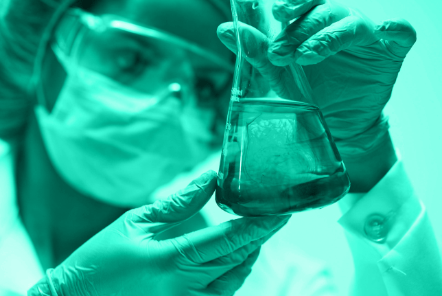 A scientist in protective gear closely examines a glass flask filled with liquid, demonstrating laboratory research or chemical analysis.