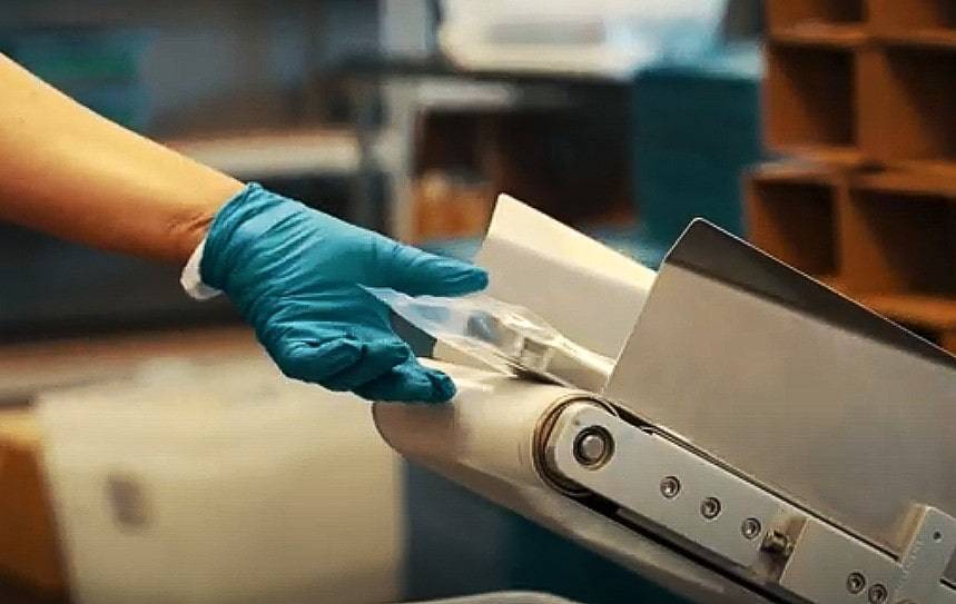 A gloved hand places an item onto a conveyor belt in a manufacturing or production setting.